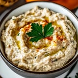 A bowl of Baba Ganoush Dip with a green herb on top.