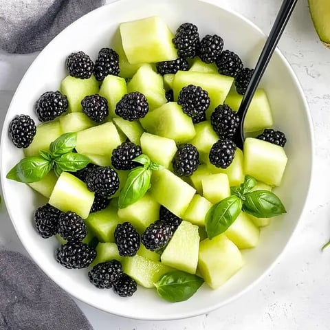 Un bol de ensalada de frutas con moras, melón verde y albahaca.